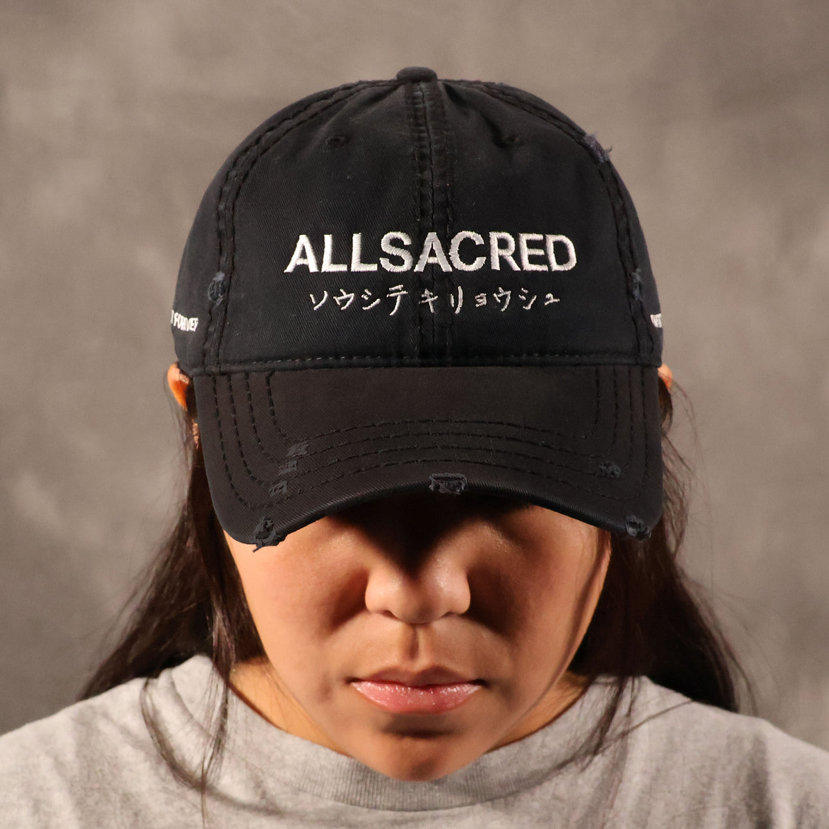 A person wearing a black distressed baseball cap with white embroidered text reading 'ALLSACRED' and Japanese characters on the front, photographed against a neutral gray background.