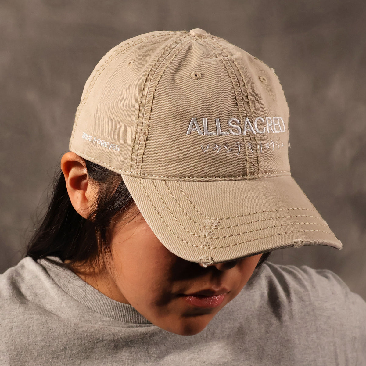 A person wearing a khaki baseball cap with white embroidered text reading 'ALLSACRED' and 'SINCE FOREVER' on a textured gray background.